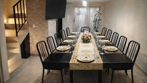 Un espace repas avec une longue table dressée pour le repas, entourée de chaises, un téléviseur fixé au mur et un escalier menant à l’étage supérieur.