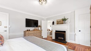 A bedroom with a bed, dresser, TV, fireplace, and a door leading to another room. - Driftwood Suite at The Royce - Coastal Luxury (Newport)
