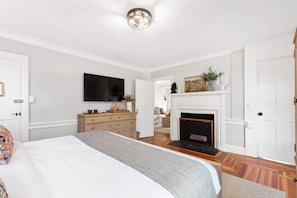 Un dormitorio con cama, cómoda, televisor, chimenea y puerta que da a otra habitación.