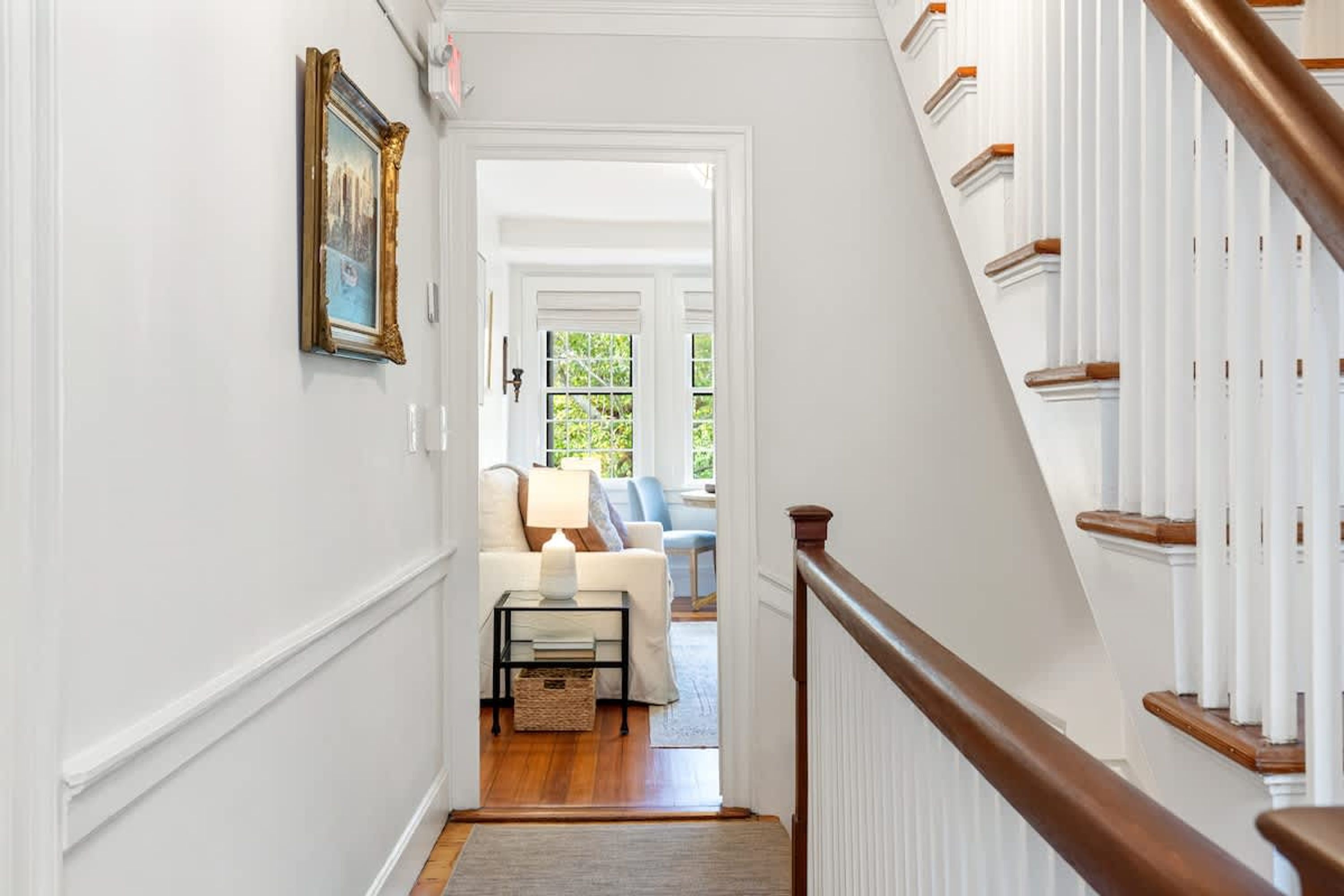 A hallway with a staircase, a framed picture on the wall, a lamp on a side table, and a view into a living room with a sofa and a lamp.