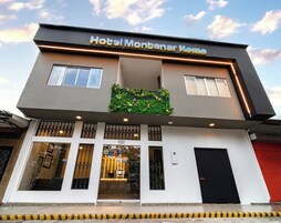 Un hotel moderno con techo plano, un cartel que dice 'Hotel Montaner Home', y un área de recepción con puerta y ventanas de vidrio.