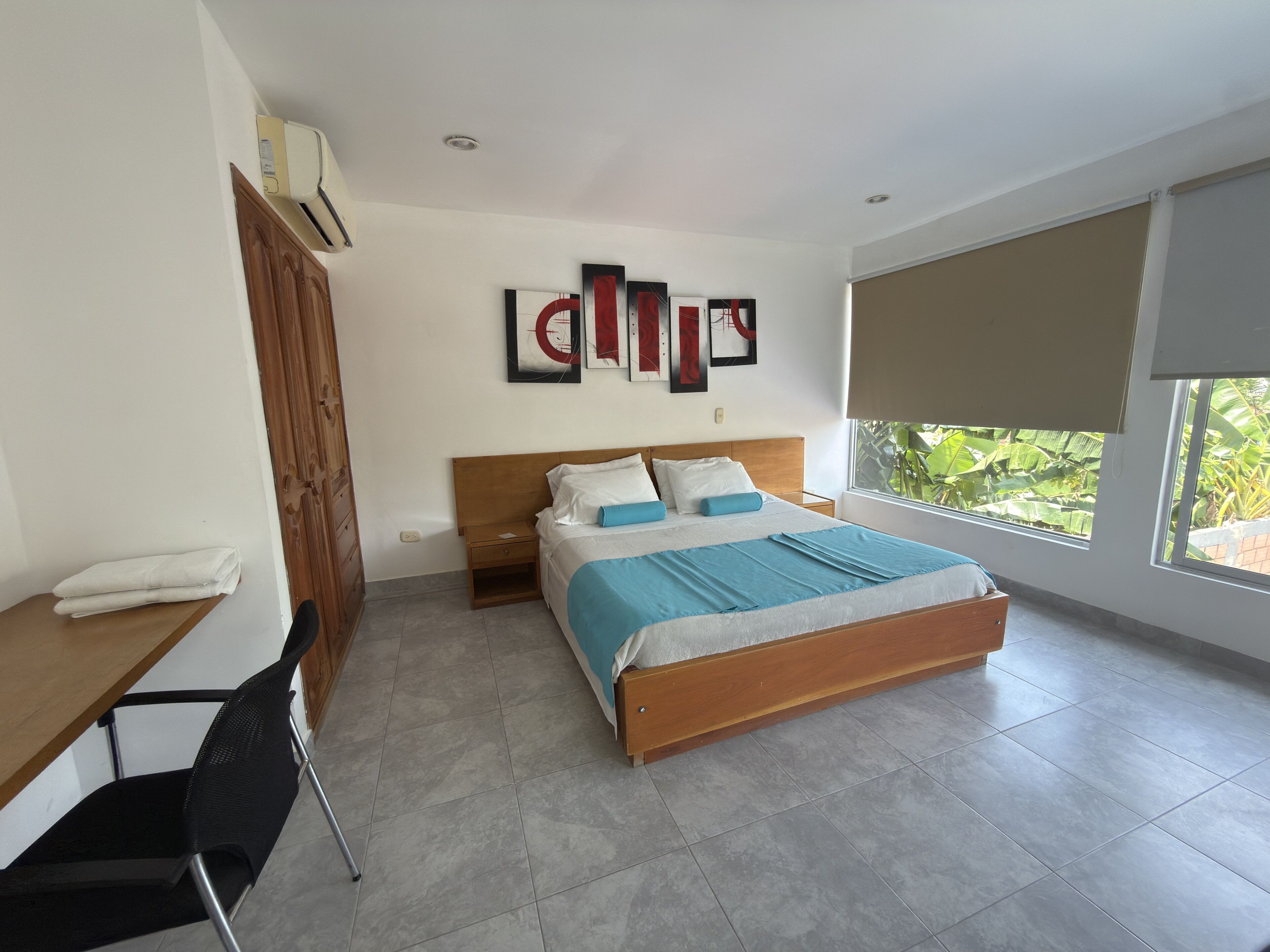 Quarto com cama, escrivaninha e cadeira. Há quadros na parede acima da cama e uma janela com vista para o verde lá fora.