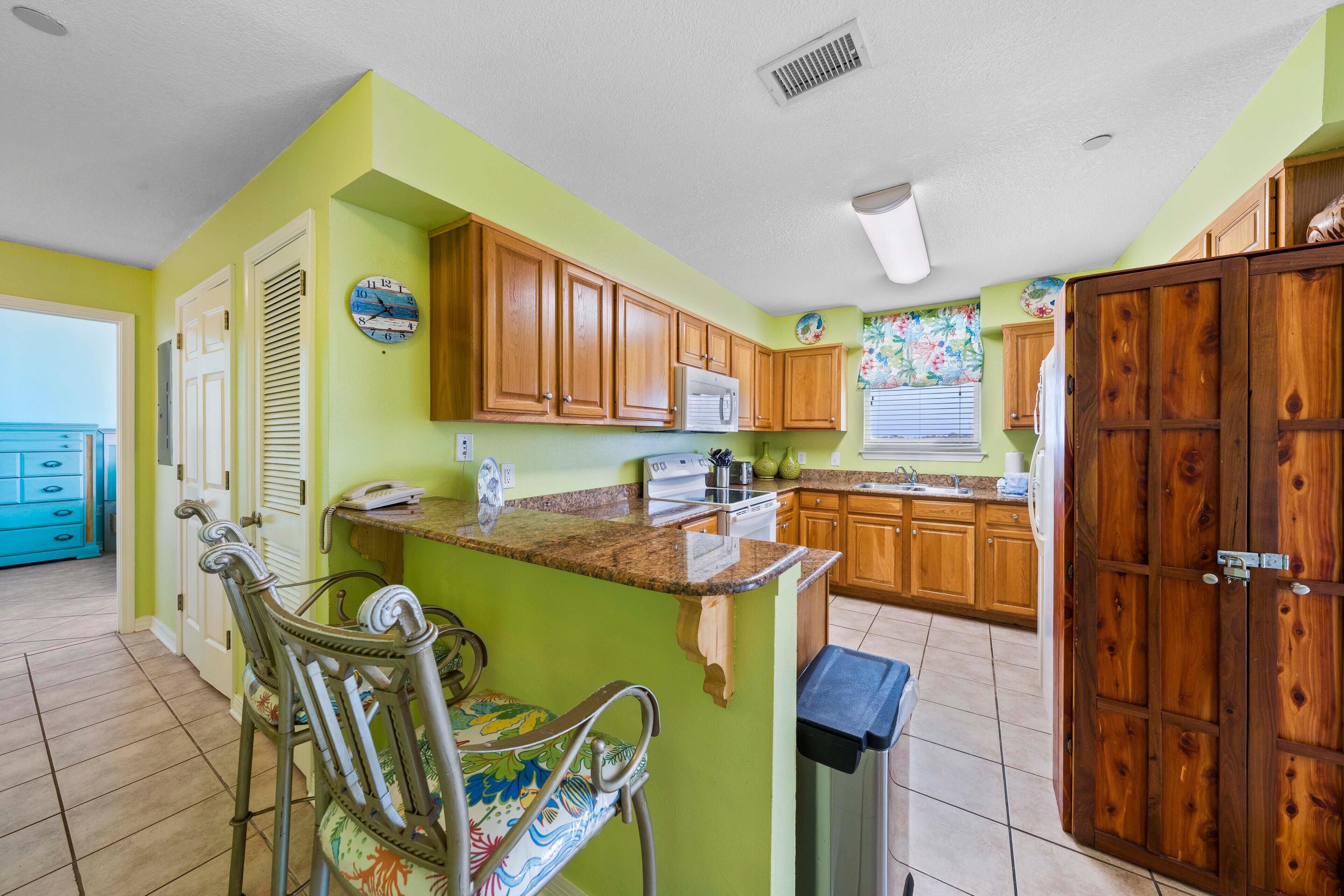 A kitchen with green walls, wooden cabinets, and a small island with a sink. There is a trash can and a chair with a colorful seat.