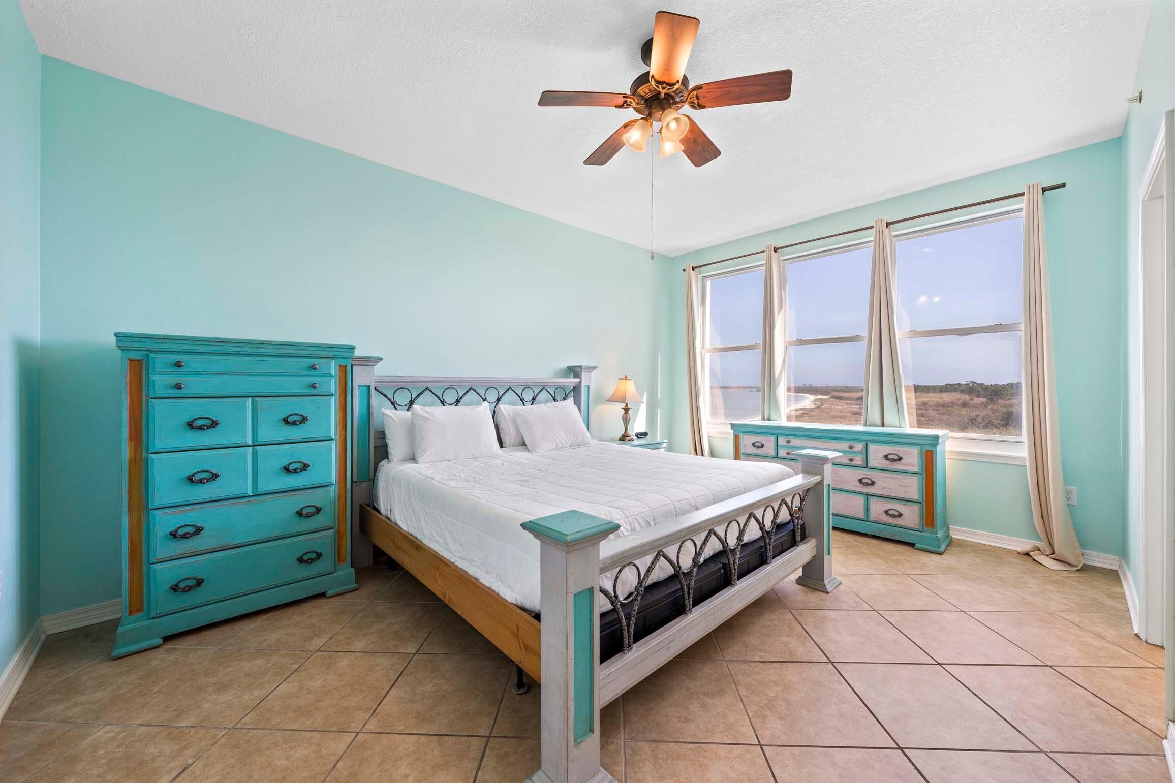 A bedroom with a bed, a dresser, a nightstand, a ceiling fan, and a window with a view.