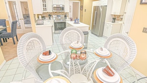 Une cuisine moderne avec des meubles blancs, des appareils en acier inoxydable et un coin repas comprenant des chaises en osier blanc et une table en verre pour quatre personnes.