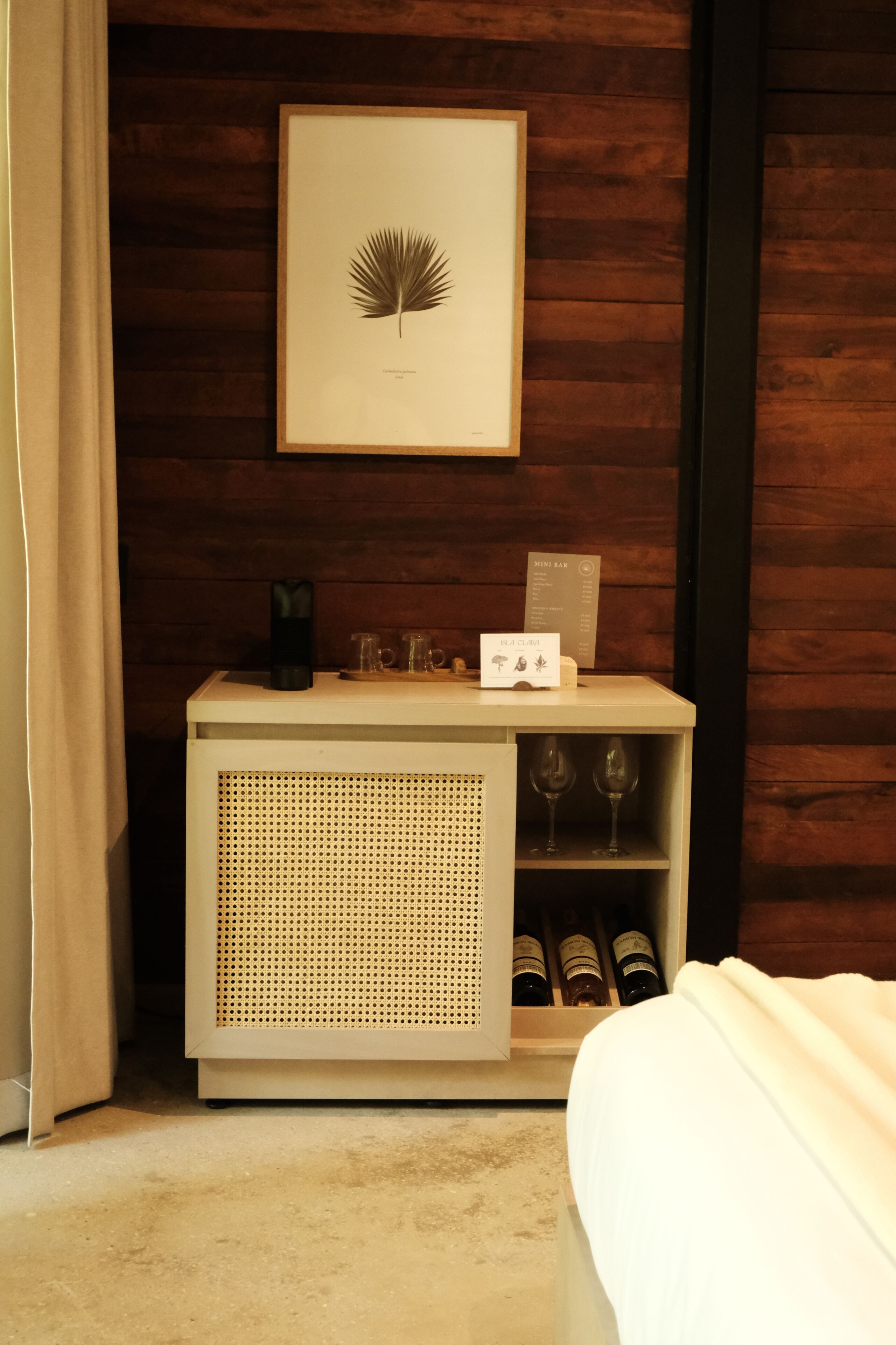 A hotel room with a wooden wall, a framed botanical print, a minibar with wine bottles, and a bed.