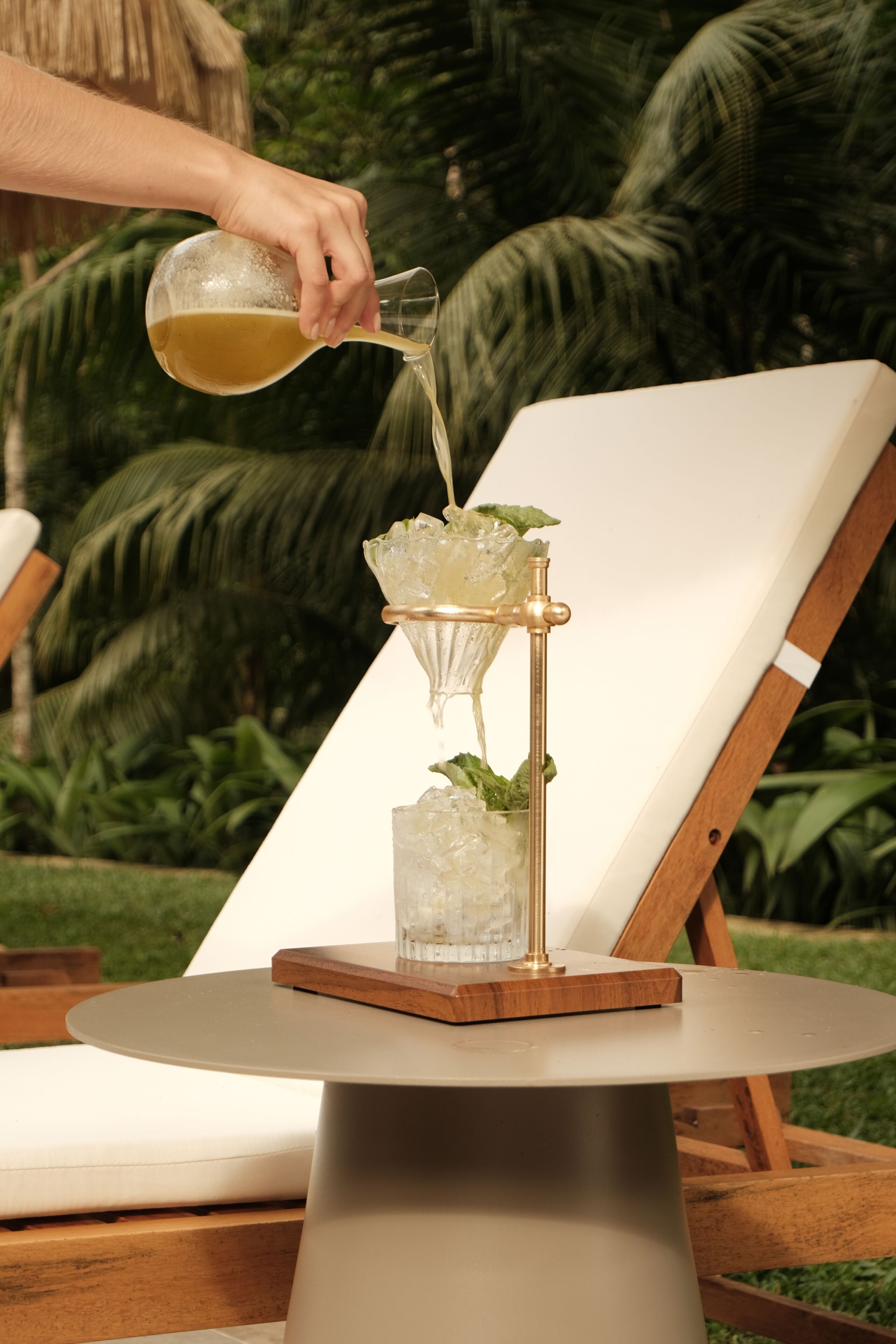 A cocktail being poured into a glass on a table with a chair in the background.