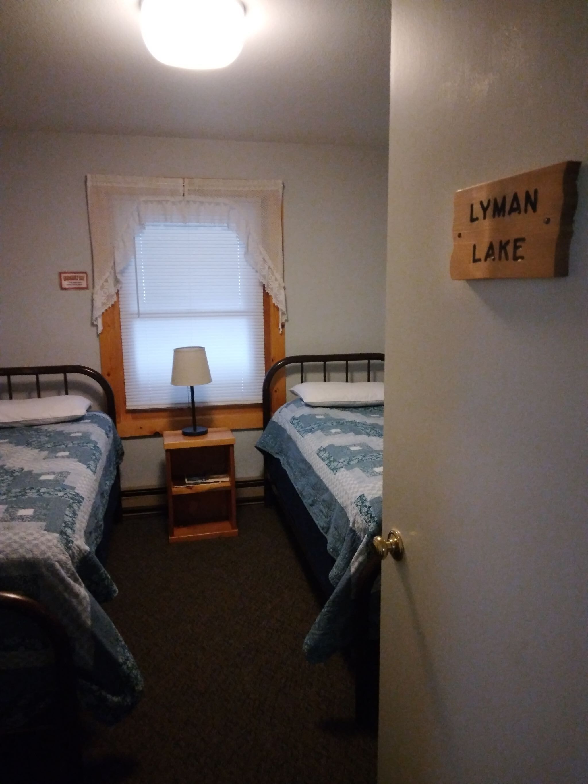 Une chambre avec deux lits, une table de chevet, une fenêtre avec des stores, une lampe et un panneau accroché au mur.
