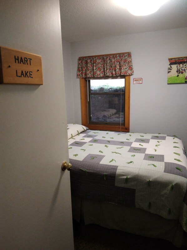 A bedroom with a bed, a window with a floral valance, a door with a sign reading "HART LAKE", and a wall-mounted picture.
