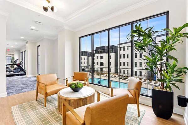 A spacious room with a large window overlooking a pool and buildings, a round wooden table with chairs, and a treadmill in the background.