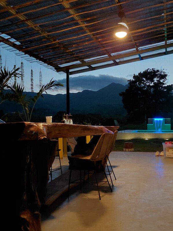 An outdoor seating area with a table and chairs, a wooden structure overhead, and a view of mountains in the background.