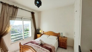 A bedroom with a bed, a nightstand, a lamp, a window with curtains, and a view of buildings outside. - Bright apartment with unobstructed view in the city center (Agadir)