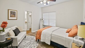 Ein Schlafzimmer mit einem Bett, einem weißen Sofa, einem kleinen Tisch mit einer Vase, einem Deckenventilator und einem Fenster mit Jalousien.