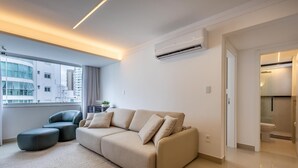 A modern living room with a beige sofa, a green armchair, and a small side table with a plant. An air conditioning unit is mounted on the wall, and a window offers a view of a building outside. - Comfort and Convenience 350 Meters From the sea in Balneario Camboriu - Rlps (Balneário Camboriú)