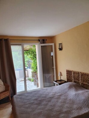 Un dormitorio con cama, una mesita de noche, una lámpara y un ventanal corredero que da a un patio con sillas y follaje.