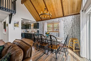 Un salon chaleureux avec un plafond en bois, un coin repas avec une table et des chaises, et une cuisine à l’arrière-plan.