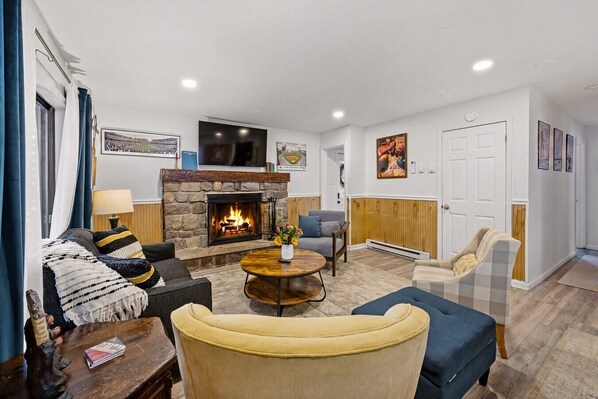 A living room with a stone fireplace, a sofa, a chair, a coffee table, and a television. - Pocono Escape | Putting Green + Sauna + Hot Tub (Lake Harmony)