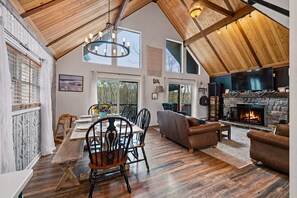 A spacious loft with a high ceiling, a dining area with a wooden table and chairs, a fireplace, and a large window overlooking greenery. - Pocono Escape | Putting Green + Sauna + Hot Tub (Lake Harmony)