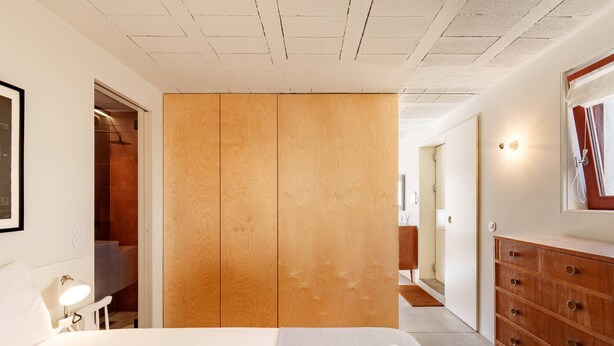 Una camera d'albergo moderna con un letto, un'armadio in legno, una cassettiera e una piccola scrivania con una lampada.