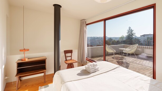 A bedroom with a bed, a wooden nightstand, a chair, a balcony with a view, and a large window. - VIVA - Lage Serviced Apartments 29 (Porto)