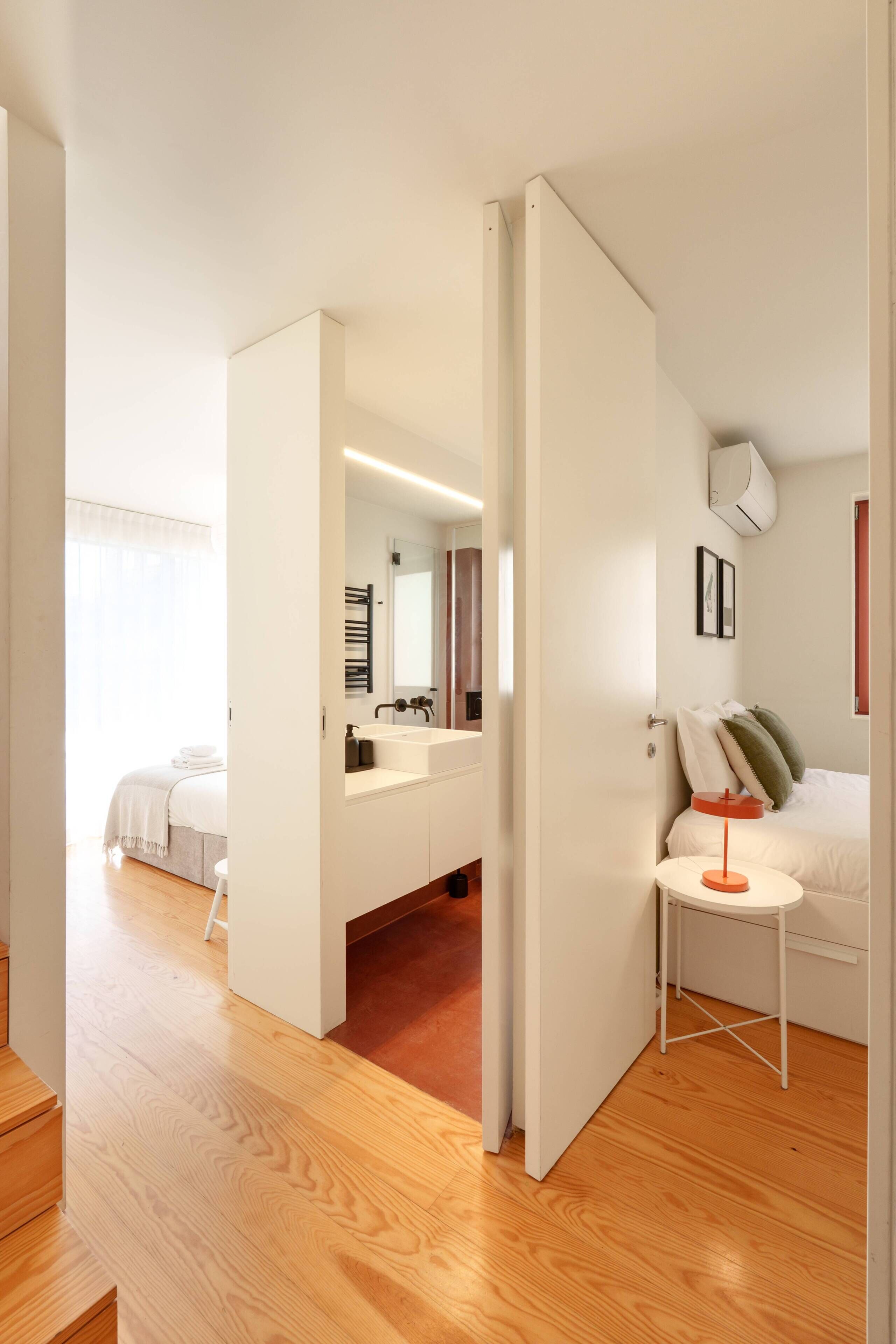 A modern bedroom with a wooden floor, a white bed, a bedside table with a lamp, a mirror, and a bathroom visible through an open door.