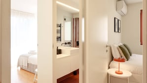 A modern bedroom with a wooden floor, a white bed, a bedside table with a lamp, a mirror, and a bathroom visible through an open door. - VIVA - Lage Serviced Apartments 29 (Porto)