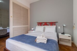 Cama bem arrumada com um cobertor azul, almofadas brancas e uma toalha dobrada. Tem uma mesinha de cabeceira com uma luminária e uma pequena mesa com um telefone.
