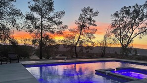 Une piscine illuminée de lumières bleues, entourée d’arbres et offrant une vue sur le coucher de soleil.