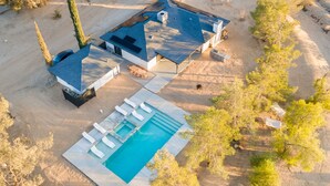 Vue aérienne d’une maison moderne dotée d’une piscine, entourée d’arbres et d’un paysage sablonneux.