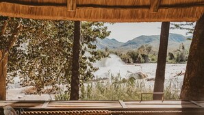 Une terrasse en bois, surmontée d’un toit de chaume, qui offre une vue sur une rivière et des collines.