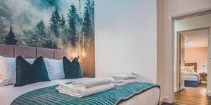 A hotel room with a bed, pillows, a quilted bedspread, a painting of a forest, and a view into another room. - Kingsley Loft – Stylish City Apartment in Central Chester | Sleeps 6 (Chester)