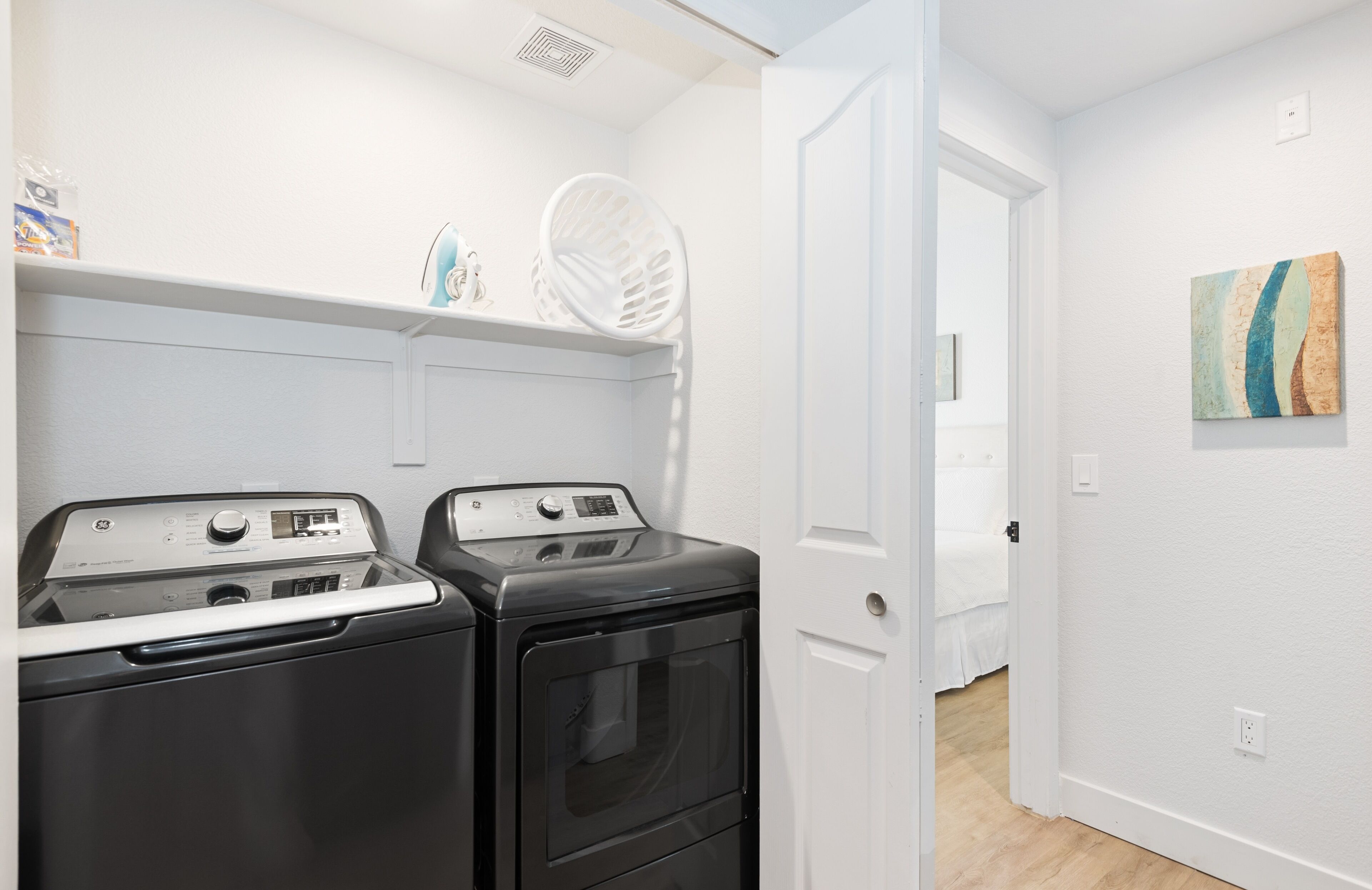 Un cuarto de lavandería con dos lavadoras y secadoras de carga frontal, un respiradero en el techo blanco y una puerta que da a otra habitación.