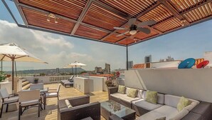 Eine Dachterrasse mit Korbmöbeln, einem Glastisch und einem weißen Sofa mit Kissen. Sonnenschirme und ein Deckenventilator sind vorhanden.