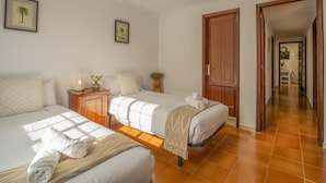 Quarto de hotel com duas camas, uma mesinha de cabeceira de madeira, um relógio fixado na parede e uma porta que dá para um corredor.