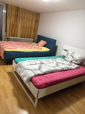 Une chambre avec deux lits, l’un doté d’une tête de lit bleue et l’autre d’une tête de lit blanche. Un radiateur se trouve sous la fenêtre, et la chambre est recouverte d’un parquet.