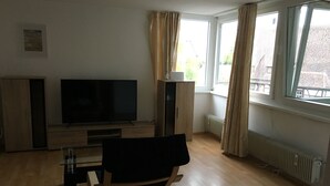 Una sala de estar con televisor, muebles de madera, radiador y ventanas amplias.