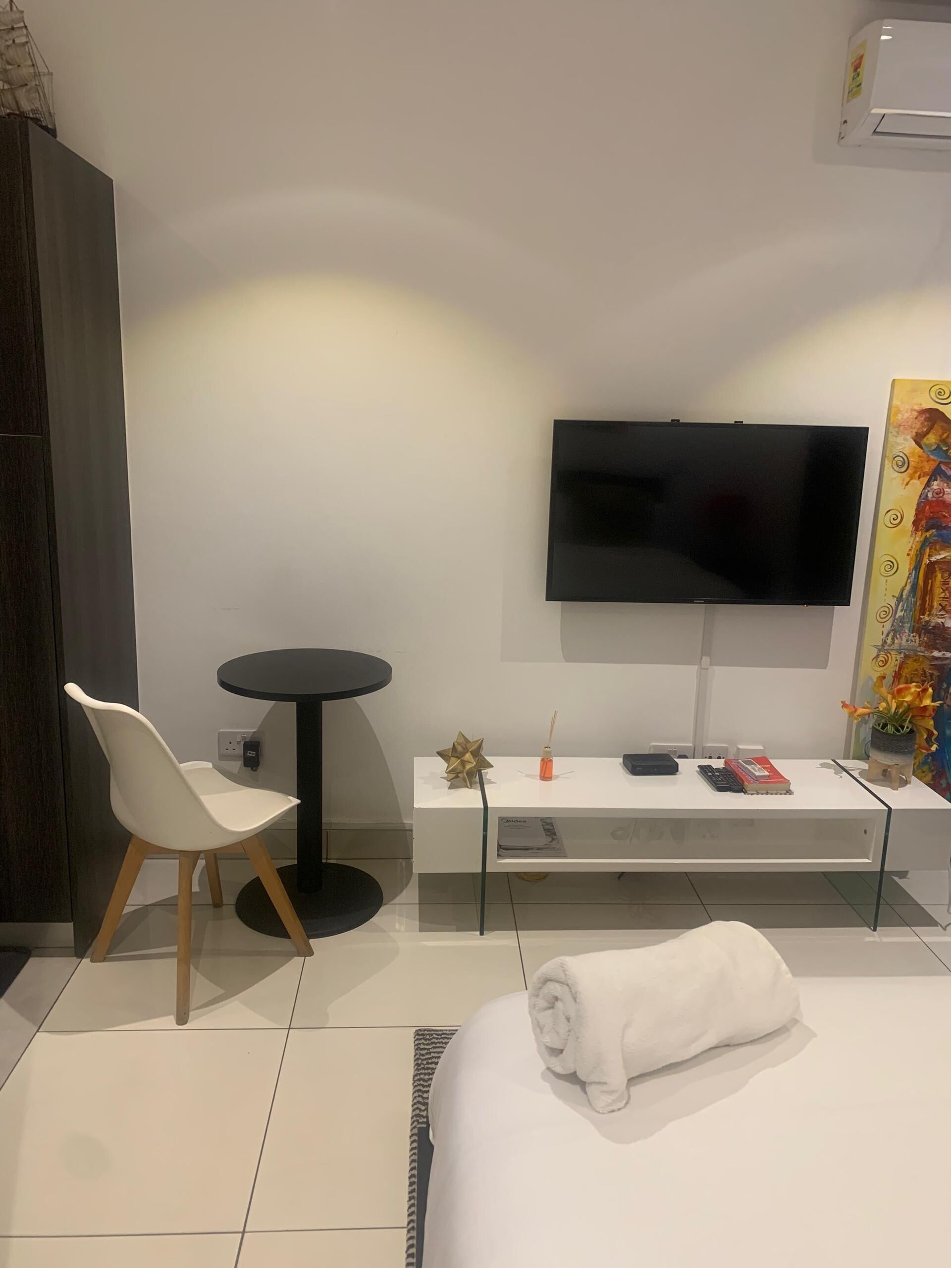 A modern hotel room with a bed, a TV mounted on the wall, a small round table with a chair, and a desk with a computer and a book.