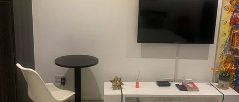 A modern hotel room with a bed, a TV mounted on the wall, a small round table with a chair, and a desk with a computer and a book.