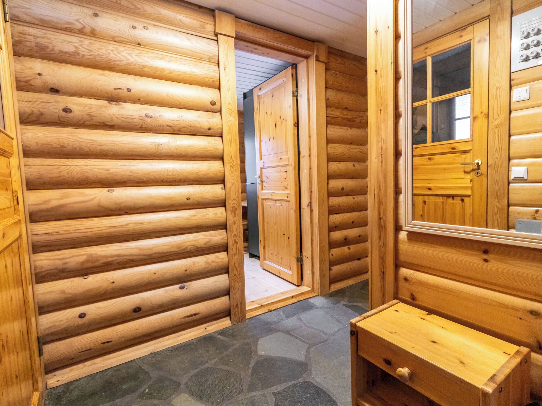 A wooden cabin interior with a stone-tiled floor, a wooden bench, and a door leading to another room.
