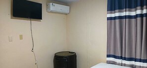 Una habitación con un televisor montado en la pared, un aire acondicionado, un cubo de basura y una cortina a rayas.