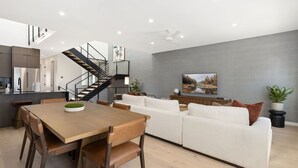 Et moderne køkken med indbygget ovn, et spiseområde med et træbord og stole, en hvid sofa og en trappe til en etage ovenover.