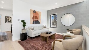 En moderne stue med sofa, sofabord og stole, et stort abstrakt maleri, et rundt spejl og en potteplante.