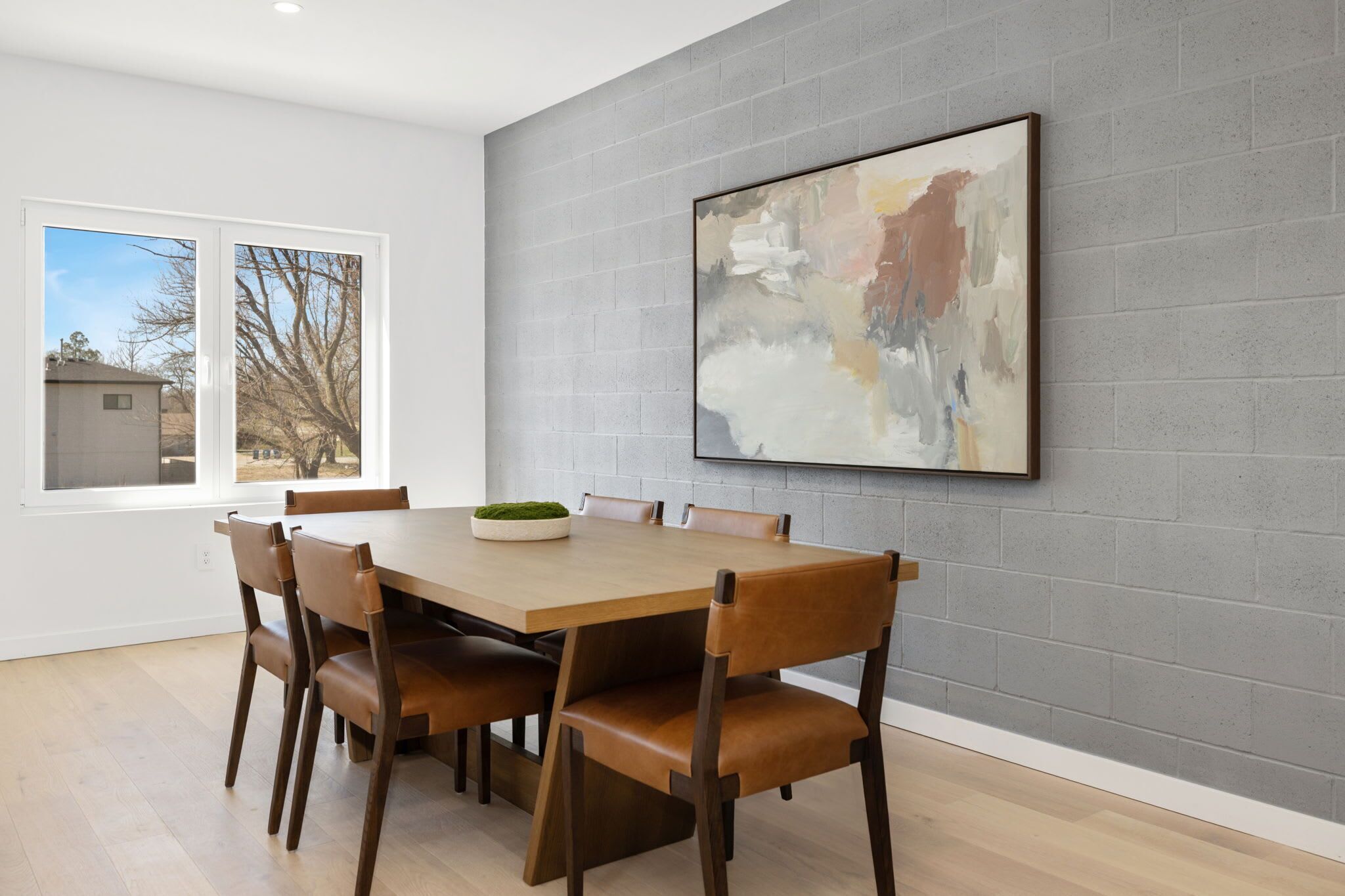 A dining area with a rectangular table, six wooden chairs, and a large abstract painting on the gray brick wall.