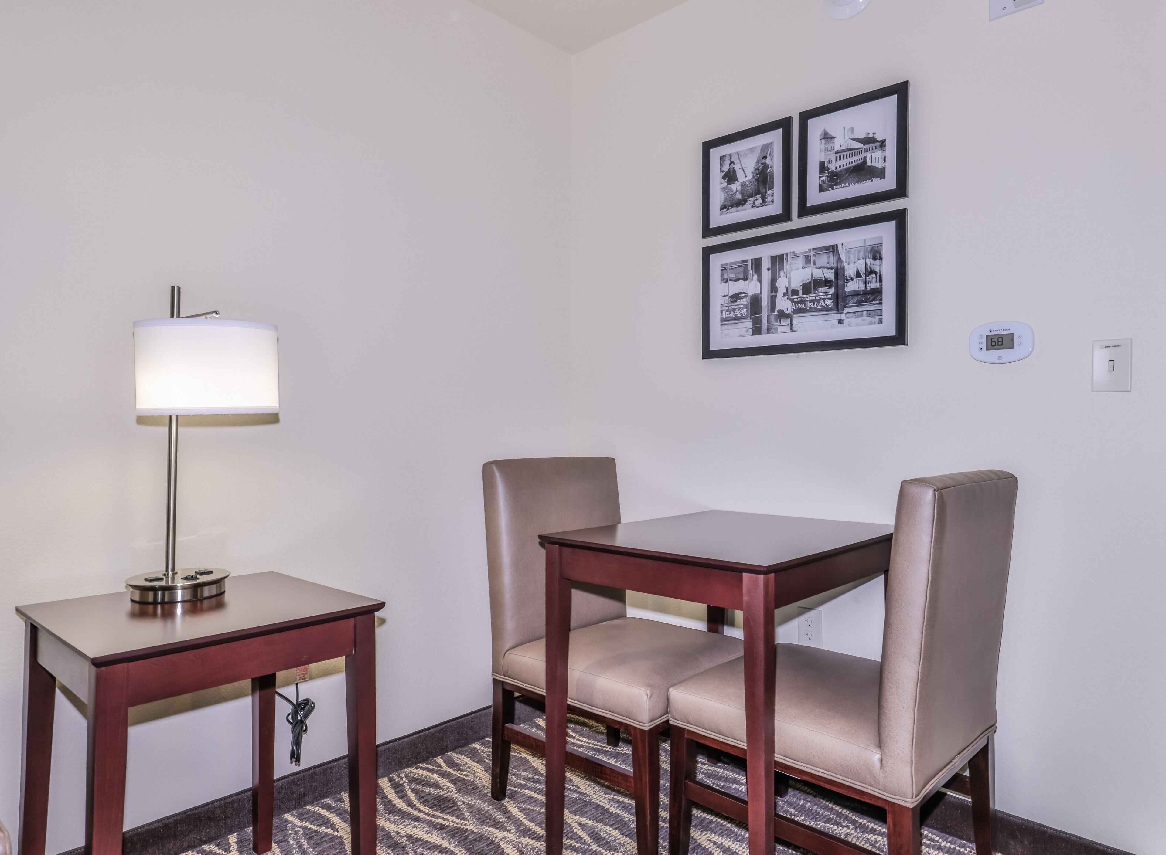 A hotel room with a wooden table and two chairs, a floor lamp, and three black-and-white photographs on the wall.