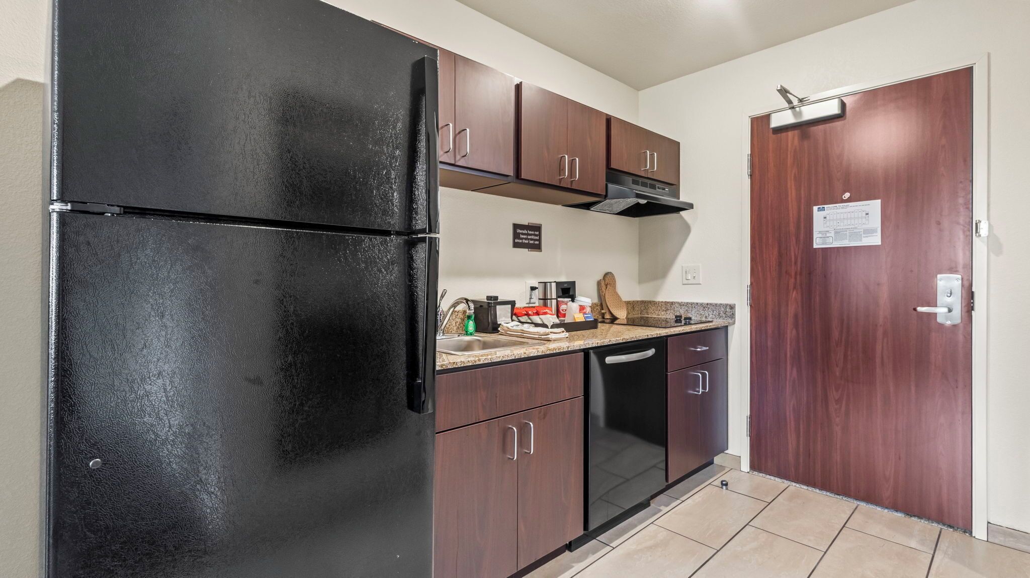 A compact kitchen with a black refrigerator, wooden cabinets, a stainless steel sink, and a door with a sign.