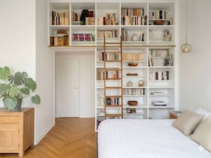 Ein Schlafzimmer mit einem großen weißen Bett, einem Holfnachtkästchen, einem Bücherregal voller Bücher und einer Topfpflanze.