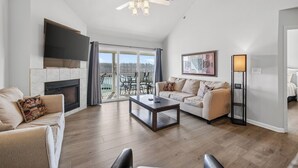 A spacious living room with a fireplace, a ceiling fan, a flat-screen TV, a sofa, a coffee table, and a large window with a view.