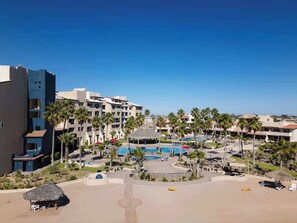 Ein Resort mit mehreren Gebäuden, einem zentralen Poolbereich, Palmen und einem Strand mit Liegestühlen.