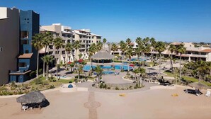 Ein Resort mit mehreren Gebäuden, einem zentralen Poolbereich, Palmen und einem Strand mit Liegestühlen.
