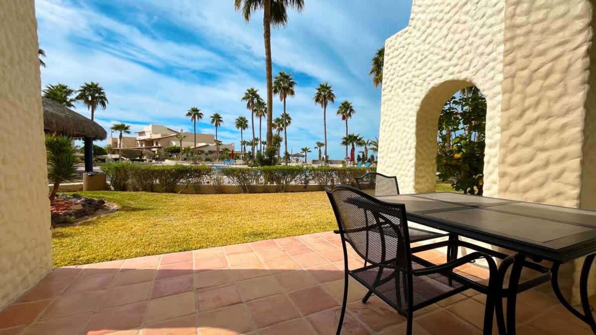A patio with a table and chairs, a pool, palm trees, and a building with a textured facade.